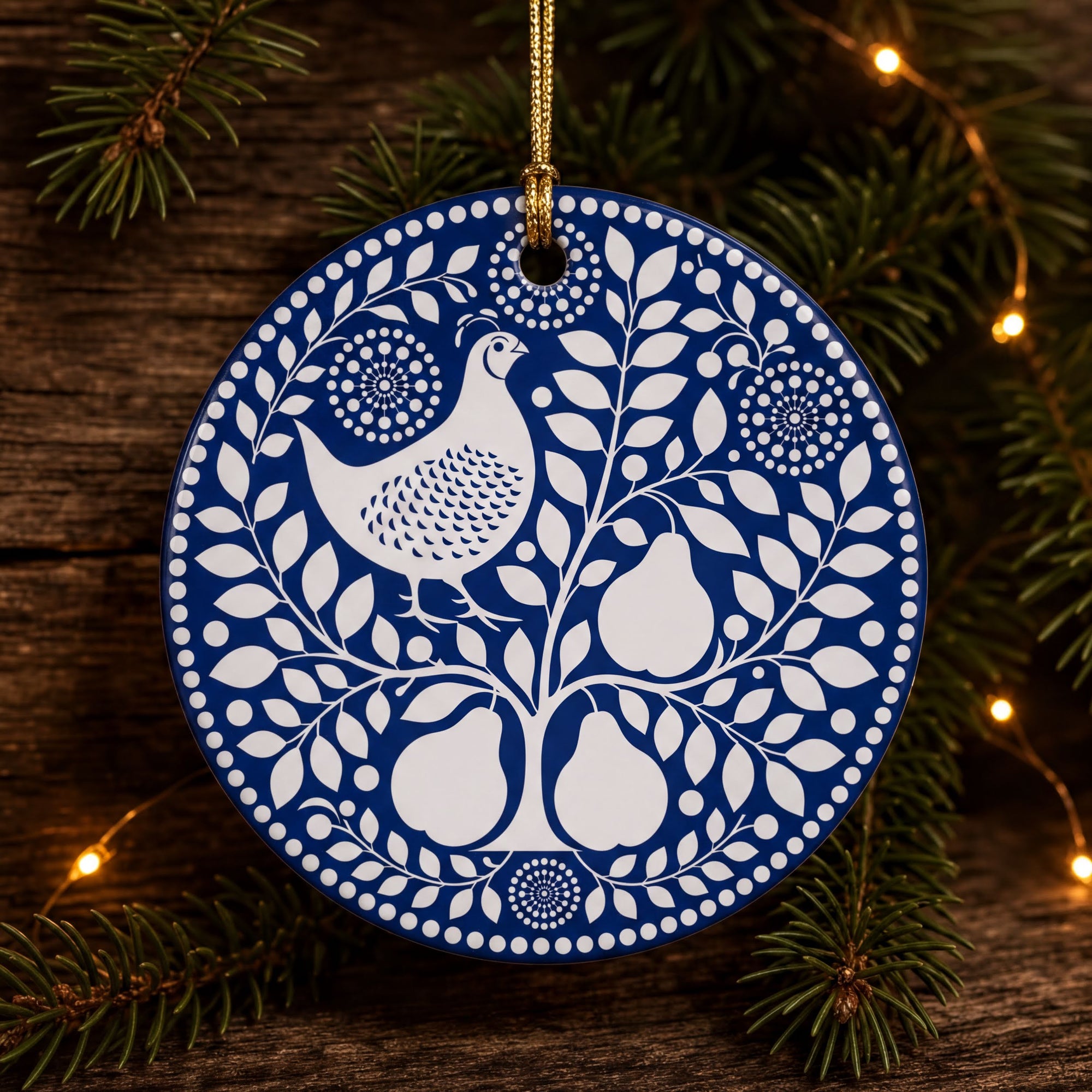 A blue and white ornament with a white bird and pears is hanging on a wooden surface, surrounded by pine branches and twinkling lights.